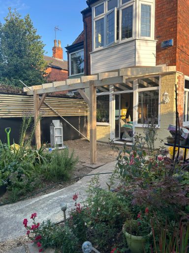 Wooden structure under construction outside a house, surrounded by plants and a garden path.